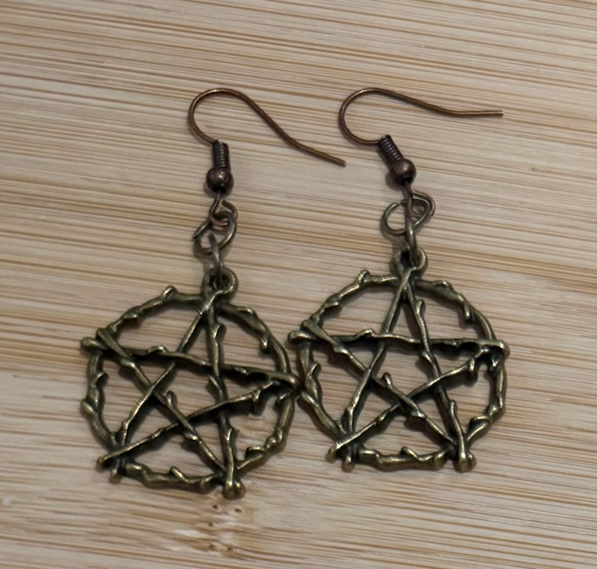 A pair of dangling earrings with an antique bronze pentagram design, displayed on a light wood grain background.