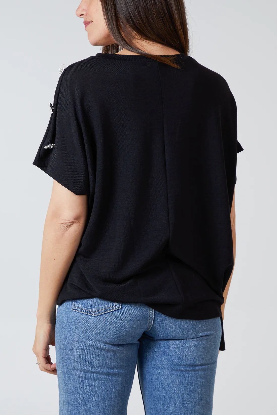 A woman shown from the back wearing a loose black short-sleeve top and blue jeans against a light gray studio background.