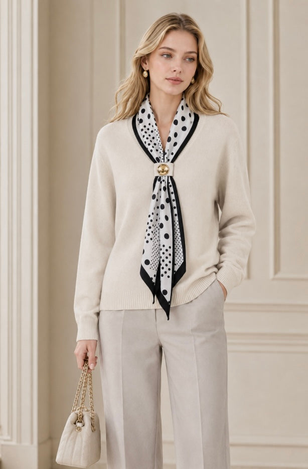 A woman models a cream knit cardigan sweater with a black and white polka dot scarf tied around the collar, styled with light gray trousers and a small cream handbag against a beige paneled wall.