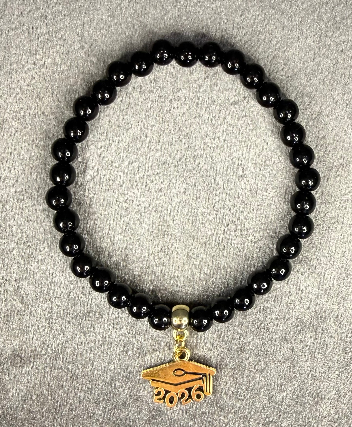 A black beaded bracelet with a gold-colored graduation cap charm featuring the year '2026' engraved on it, displayed on a textured gray fabric background.