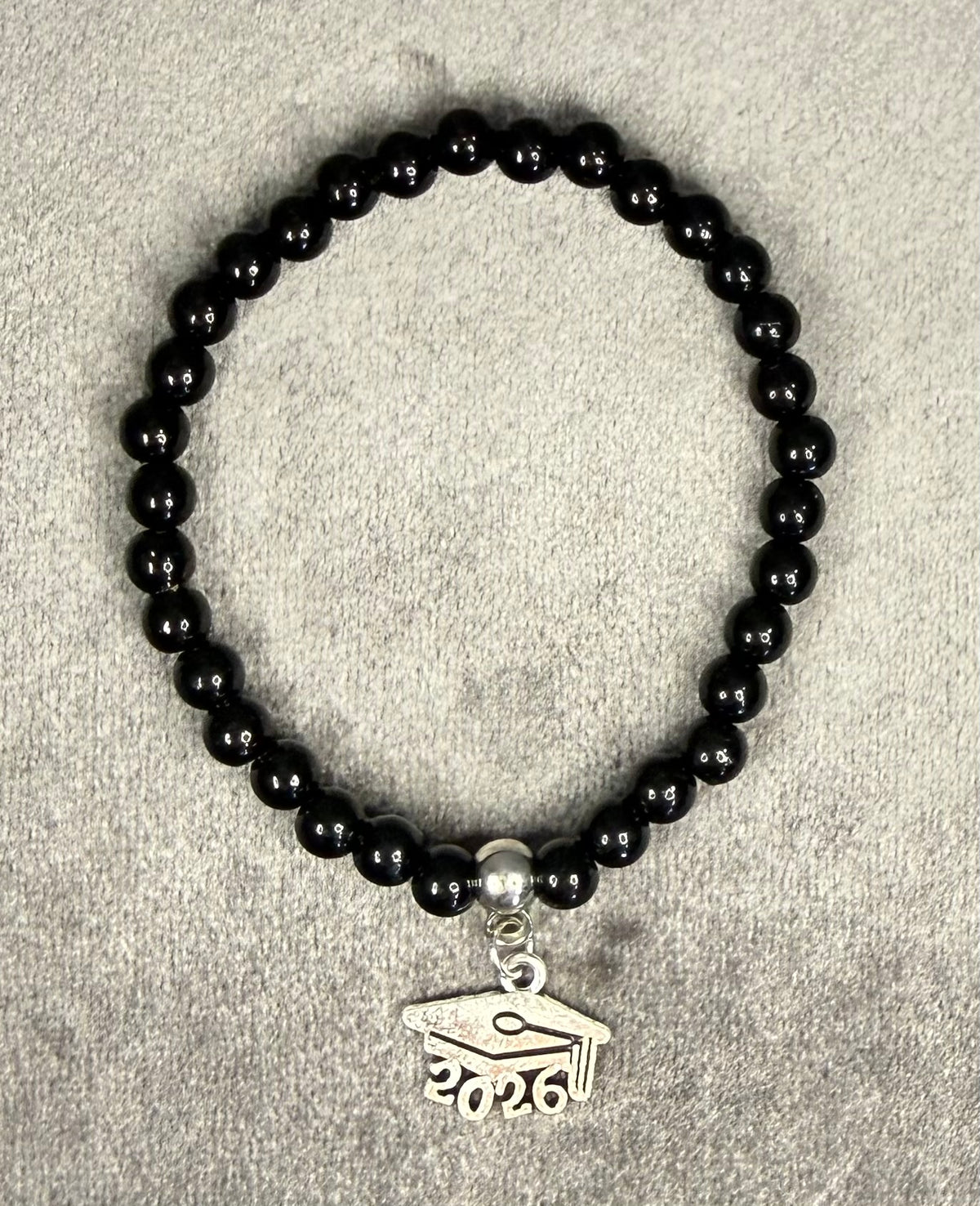 A beaded stretch bracelet made of glossy black round beads, featuring a silver-toned charm shaped like a graduation cap with the year 2026 engraved on it, displayed on a textured gray background.