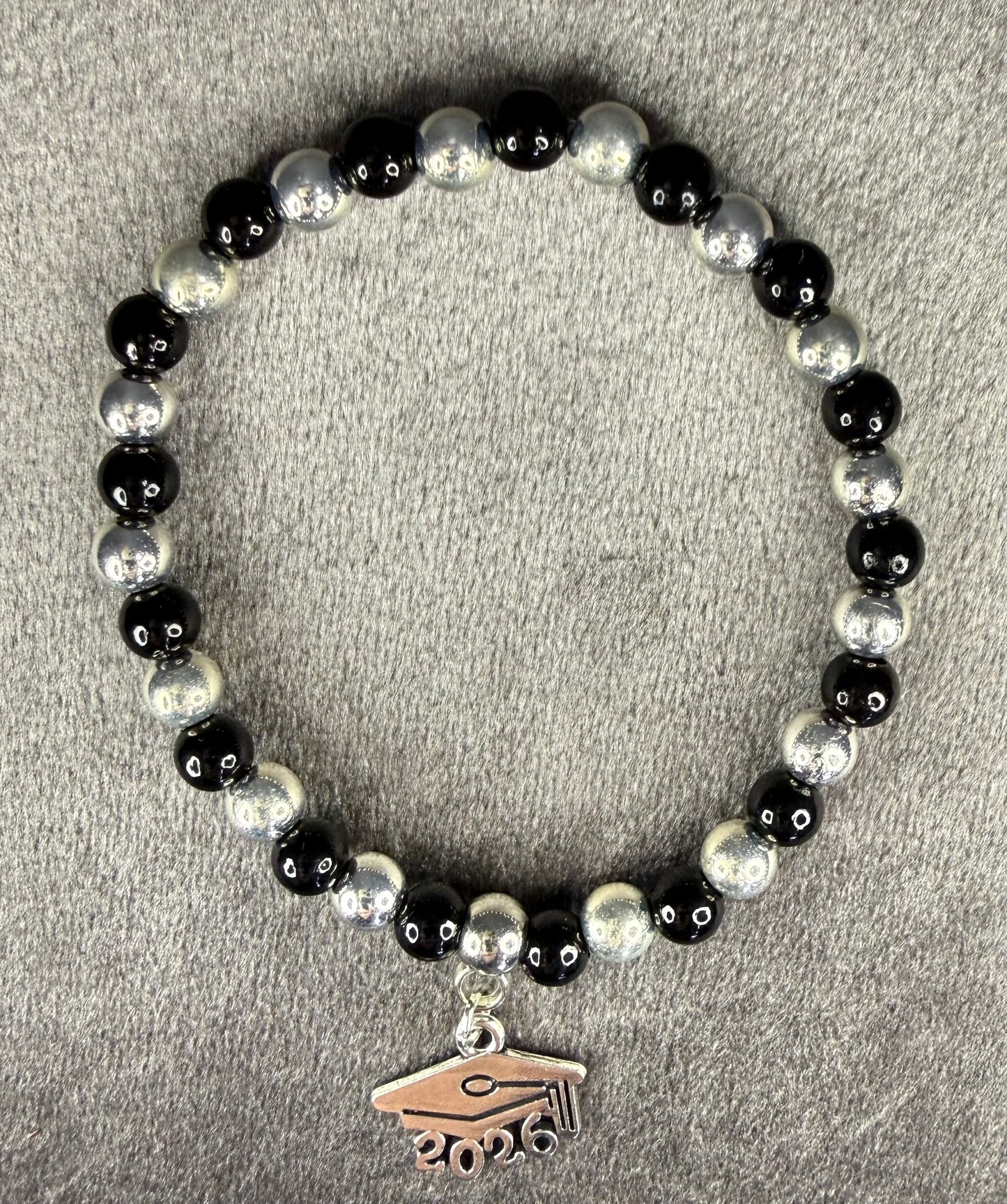 A beaded stretch bracelet featuring alternating glossy black and textured silver-gray beads, with a small silver-toned graduation cap charm engraved with the year 2026, displayed on a neutral gray textured background.