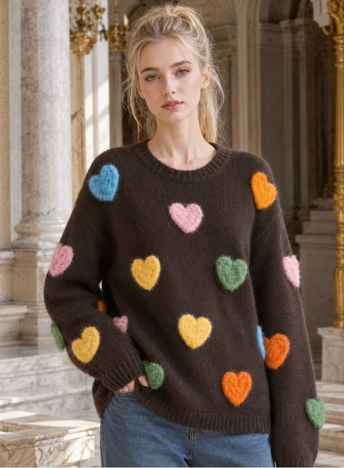 A woman wearing a dark brown knit sweater with colorful fuzzy heart patches in pink, blue, yellow, green, and orange, standing in a grand classical interior with stone columns.
