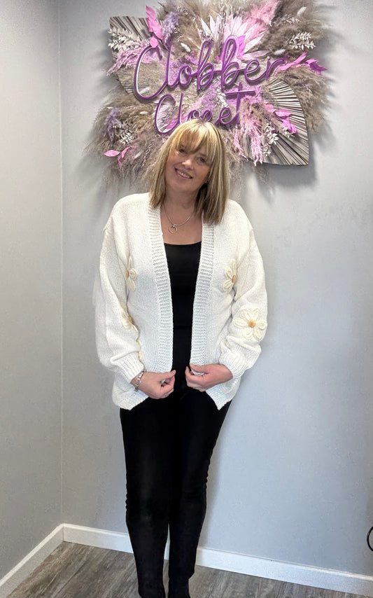 Woman wearing an Edge to Edge Open Front 3D Daisy Flower Cardiganstanding in front of a decorative wall sign with 'Clobber Clozet' on it.