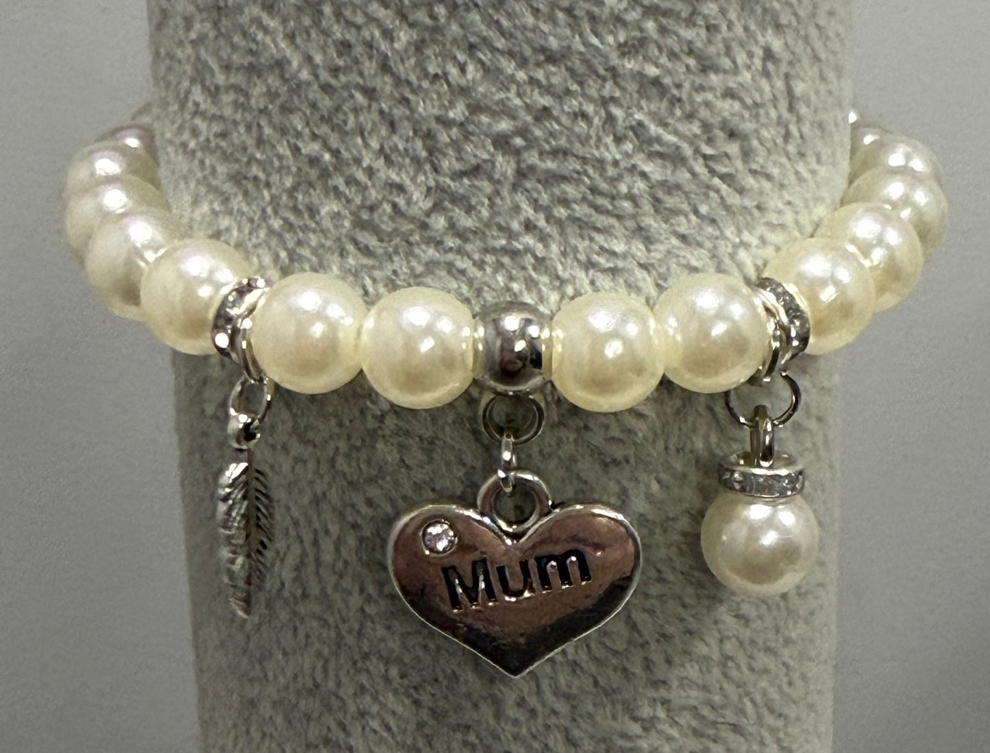 A beaded bracelet with cream-colored faux pearls, silver-toned spacer beads, and three charms: a feather, a heart engraved with 'Mum', and a small pearl with a rhinestone cap, displayed on a gray textured stand.
