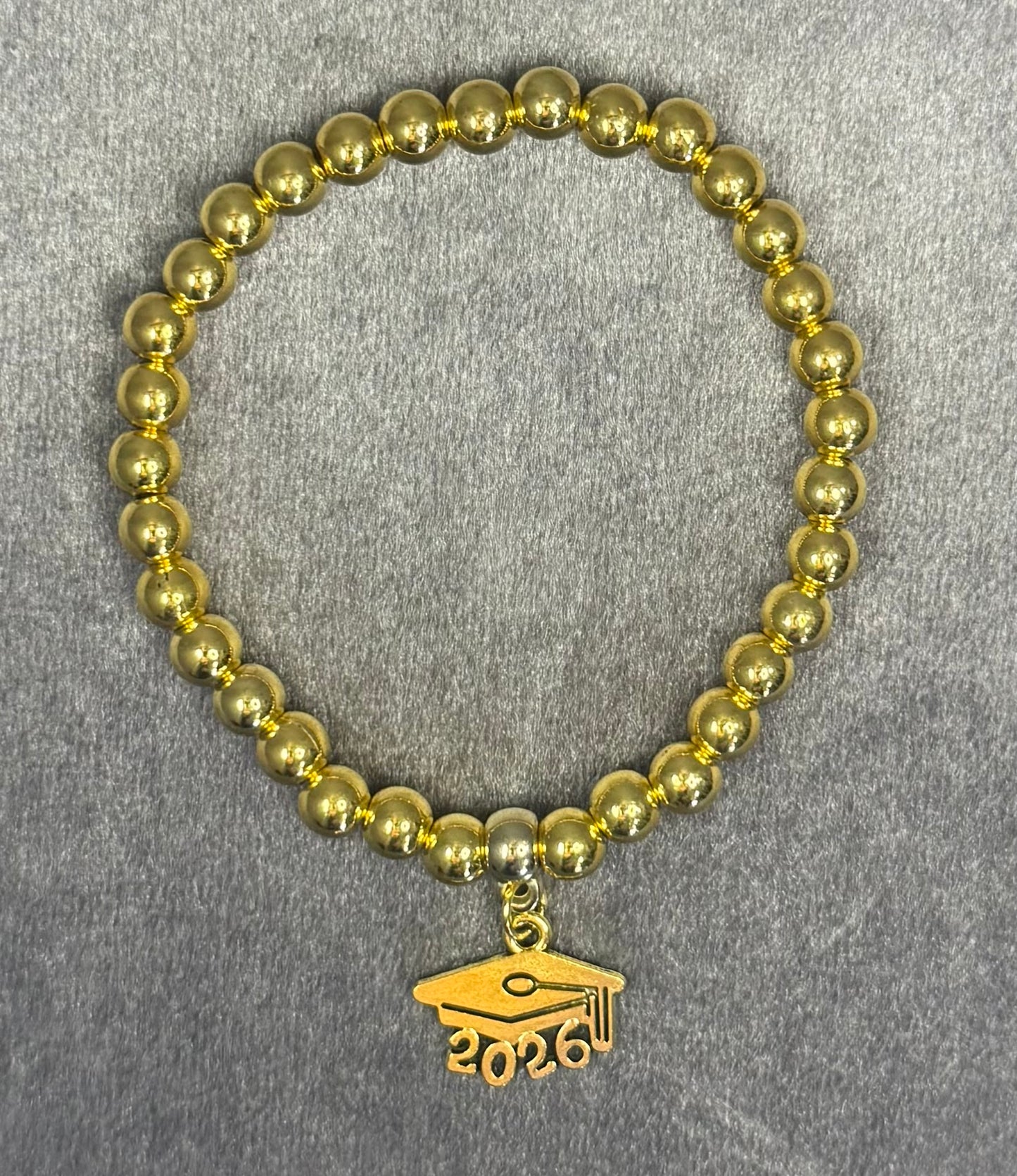 A gold beaded bracelet with a graduation cap charm engraved with '2026', displayed on a textured gray background.