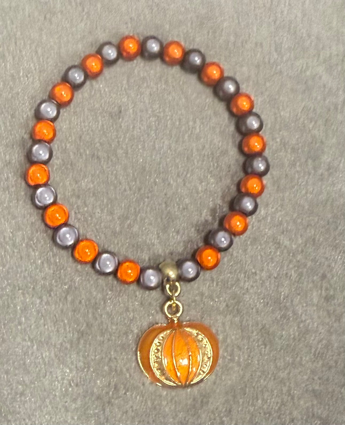 A beaded stretch bracelet featuring alternating orange and gray beads, with a small gold-toned pumpkin charm hanging from the center, displayed on a textured gray background.
