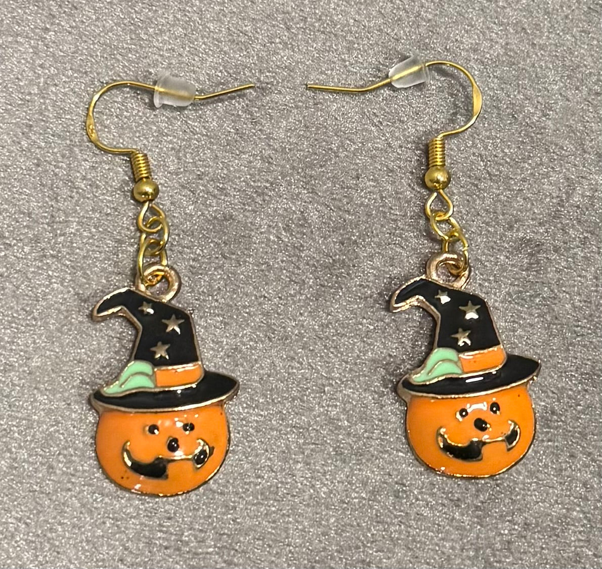 A pair of dangling earrings featuring orange pumpkin faces wearing black witch hats with mint green trim and silver star details, shown on a textured gray background.