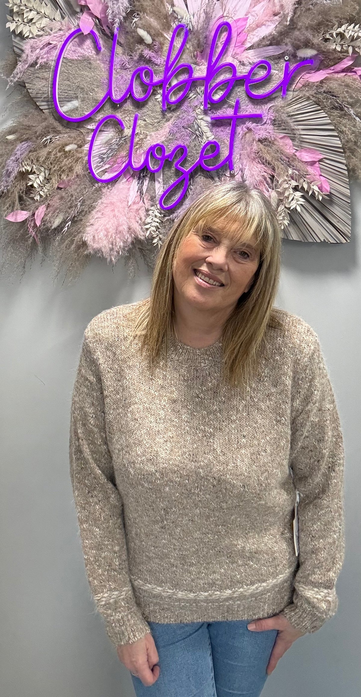 A woman wearing a beige and brown heathered knit sweater stands in front of a gray wall with a decorative dried floral arrangement and a purple 'Clobber Closet' sign. She is also wearing blue jeans.