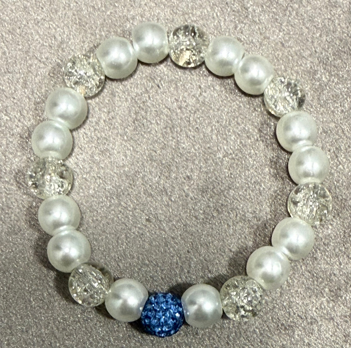 A beaded stretch bracelet featuring white faux pearls, clear faceted crystal beads, and a single prominent blue rhinestone accent bead, displayed on a textured gray background.