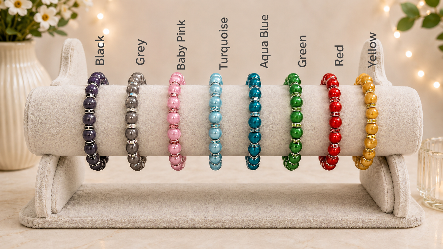 A display stand holding multiple beaded bracelets in different colors, labeled from left to right as Black, Grey, Baby Pink, Turquoise, Aqua Blue, Green, Red, and Yellow. The bracelets are arranged on a light beige velvet jewelry display against a softly blurred warm indoor background with string lights.