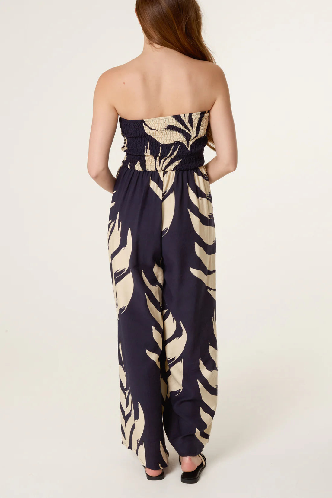 Back view of Woman wearing a strapless Navy Bandeau Tassel Leaf Print Jumpsuit with leaf pattern on a white background
