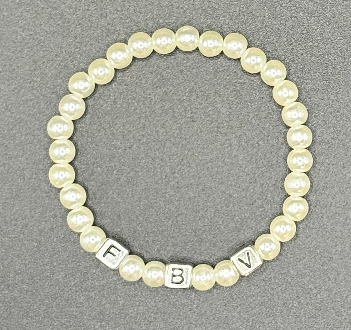 A beaded stretch bracelet featuring cream-colored faux pearls and silver letter initialcubes 'F B V', displayed on a textured gray background.