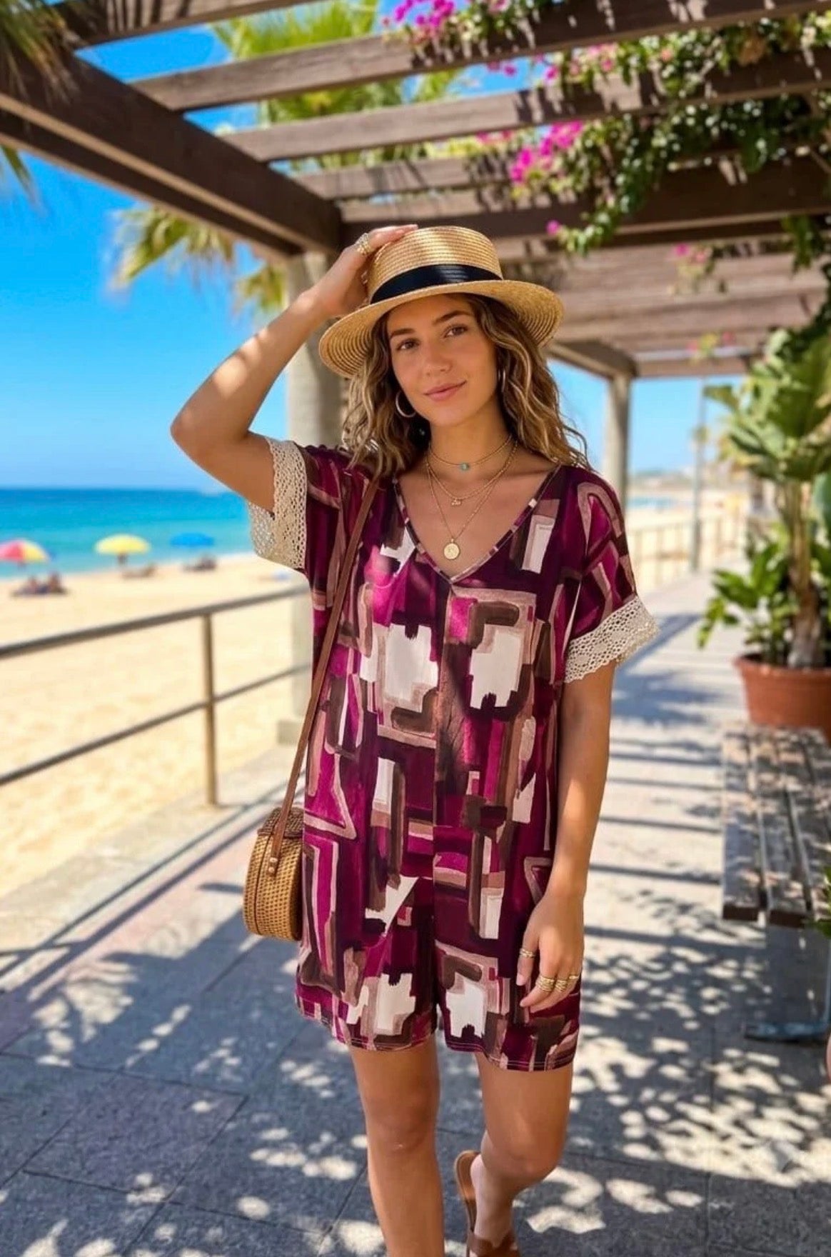 A woman wearing a short-sleeve patterned romper with a straw hat stands on a beach boardwalk. The romper has a geometric print in deep purple, burgundy, and cream, with white lace trim on the sleeves.