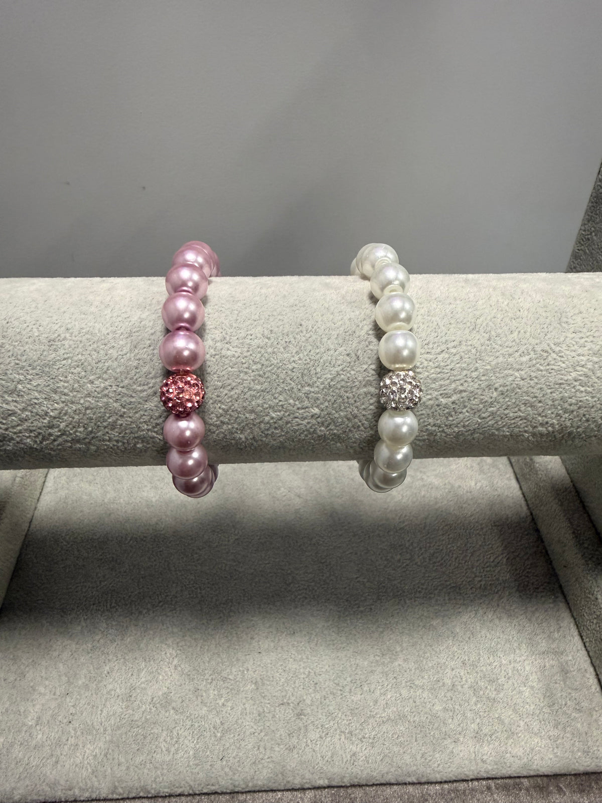 Two beaded stretch bracelets displayed on a gray jewelry stand. The top bracelet is white with a central clear rhinestone cluster, and the bottom bracelet is light pink with a central pink rhinestone cluster.