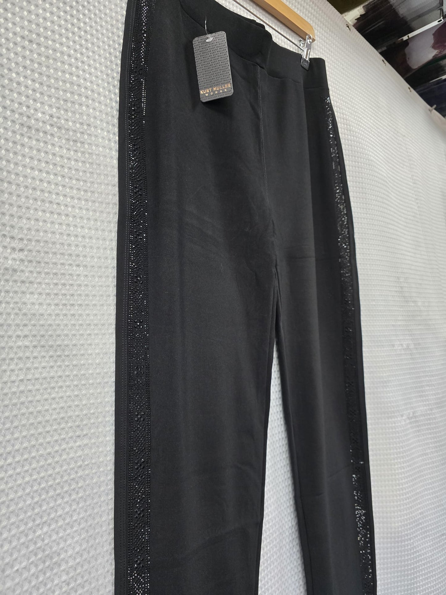 Black trousers with black diamantes down the sides hanging on a rack against a textured white wall.