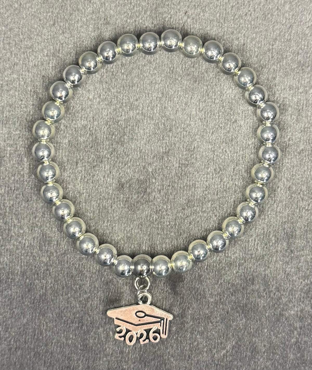 A silver beaded stretch bracelet with a graduation cap charm engraved with '2026', displayed on a textured gray background.