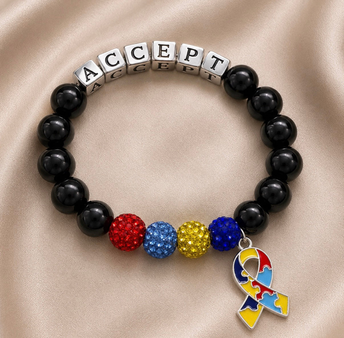 A beaded bracelet with black round beads, silver cube beads spelling 'ACCEPT', three textured multicolor beads in red, blue, and gold, and a puzzle-piece awareness ribbon charm, displayed on a beige satin background.
