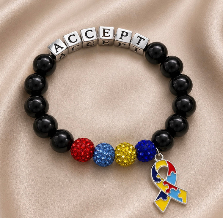 A beaded bracelet with black round beads, silver cube beads spelling 'ACCEPT', three textured multicolor beads in red, blue, and gold, and a puzzle-piece awareness ribbon charm, displayed on a beige satin background.