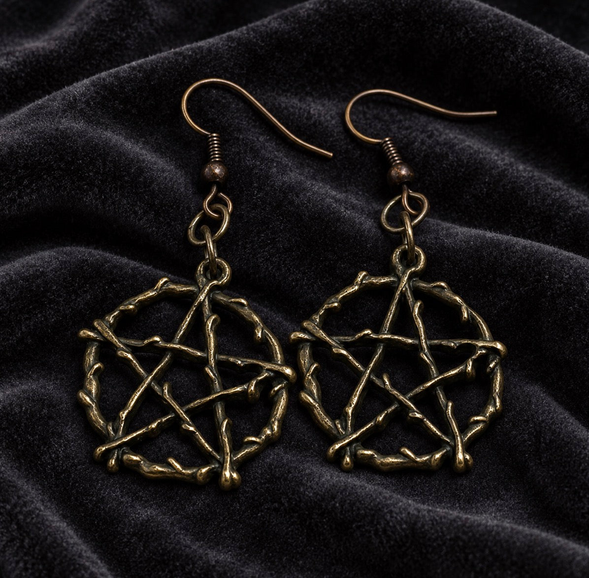 A pair of dangling earrings with twisted metal wire pentagrams inside hexagonal rings, shown on a dark textured velvet background.