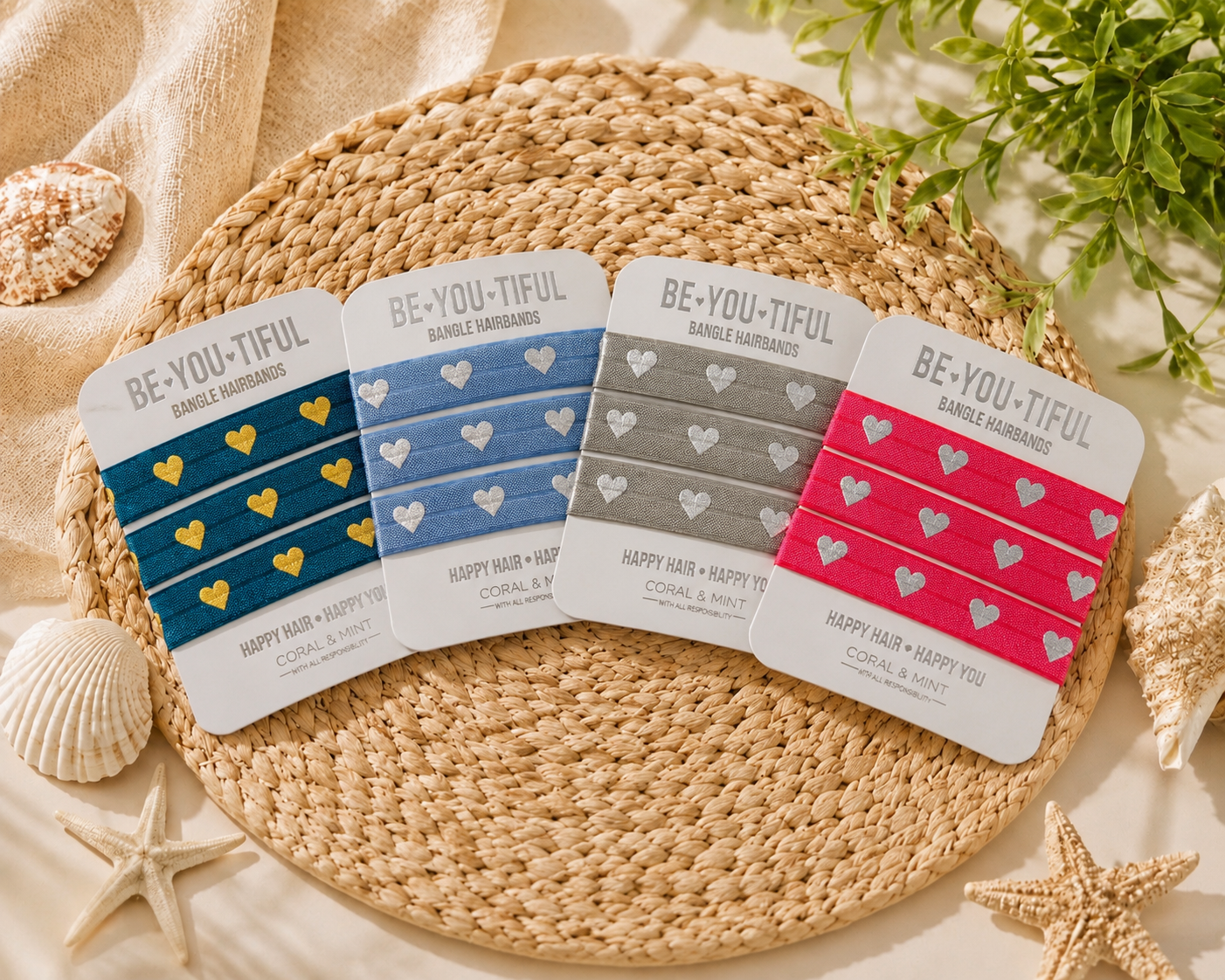 Four packaged hair wraps arranged on a woven round mat with beach-themed props including seashells, starfish, and greenery. The packages are labeled BE-YOU-TIFUL and Happy Hair + Nails, shown in blue, gray, and pink variants with heart patterns.
