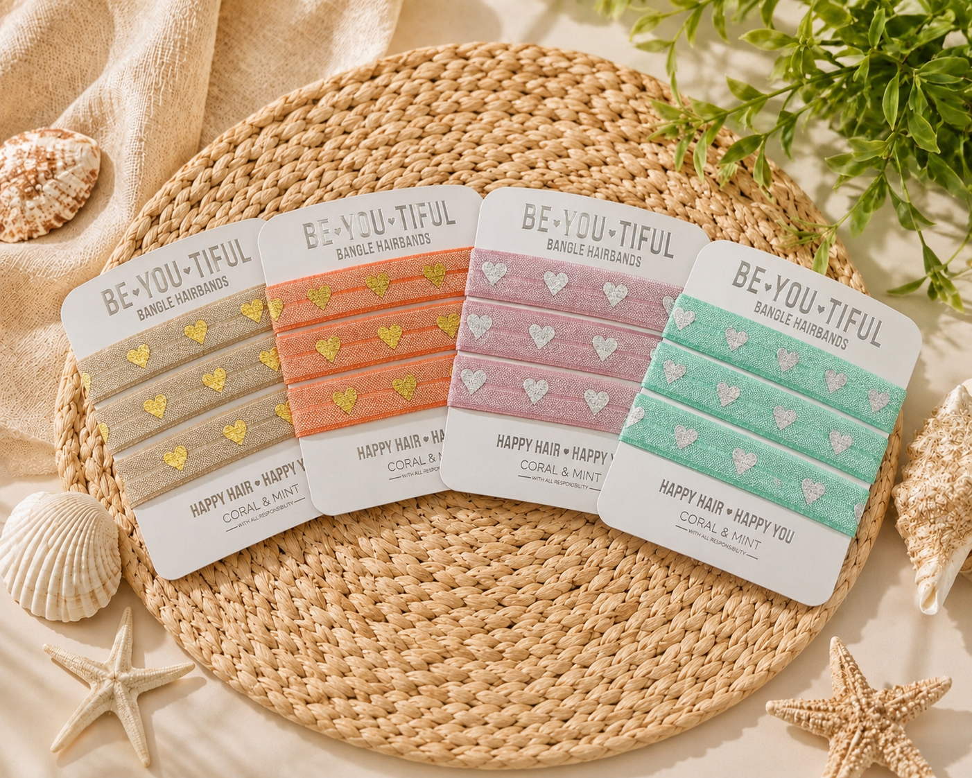 Four packaged hair elastic bands arranged on a round woven placemat with beach-themed props. The bands come in beige, coral-pink, dusty pink, and mint green, each with matching pastel hearts and gold or silver lettering.