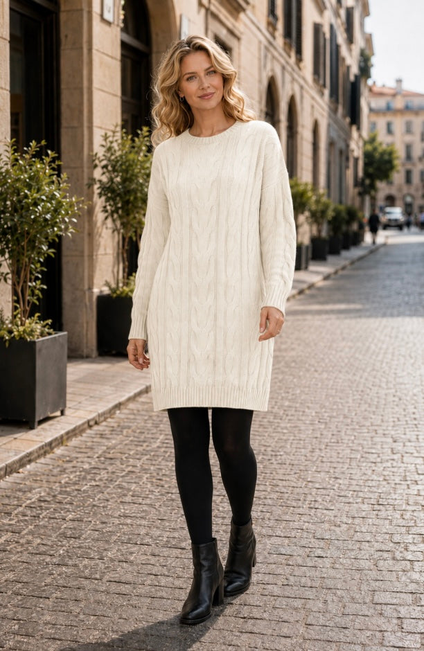 A woman stands on a cobblestone street wearing a cream cable-knit long-sleeve sweater dress, black tights, and black ankle boots.