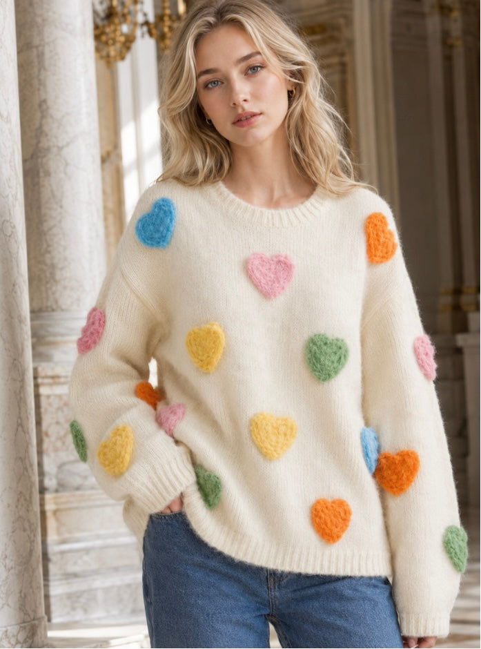 A woman wearing a cream-colored fuzzy knit sweater with colorful textured heart patches in pink, blue, yellow, green, and orange, paired with blue jeans, standing in a grand classical interior.