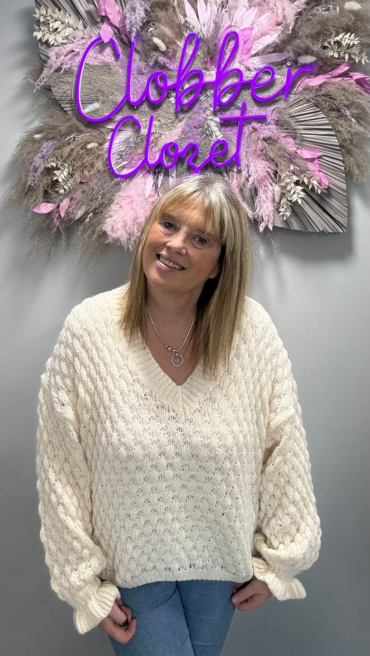 A woman wearing a chunky cream cable-knit V-neck sweater and blue jeans, standing in front of a gray wall with a decorative pink and brown wall hanging and purple 'Clobber Closet' text.