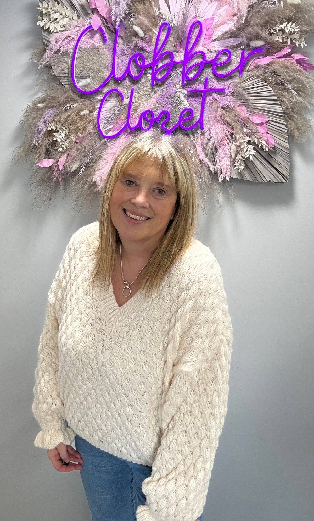 A woman with blonde hair wearing a chunky cream cable-knit sweater and blue jeans, standing in front of a gray wall with a decorative dried floral arrangement and a purple neon sign that reads 'Clobber Cloret'.