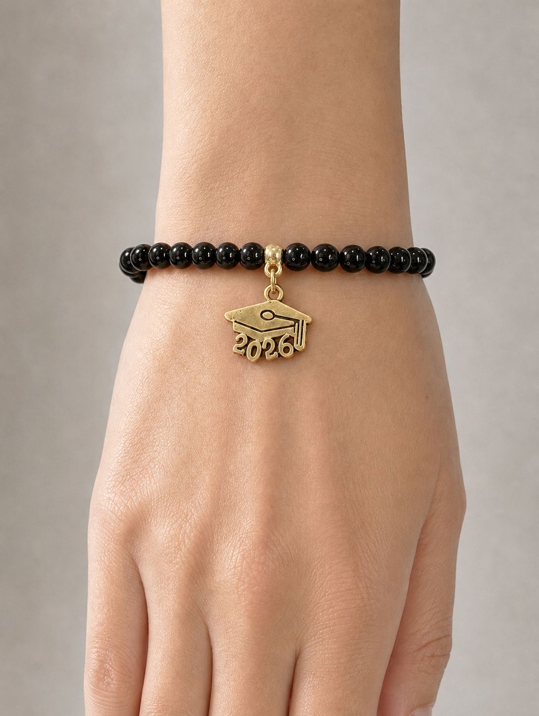 A close-up of a person's wrist wearing a black beaded bracelet with a gold graduation cap charm engraved with '2026'.