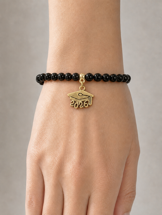 A close-up of a person's wrist wearing a black beaded bracelet with a gold graduation cap charm engraved with '2026'.