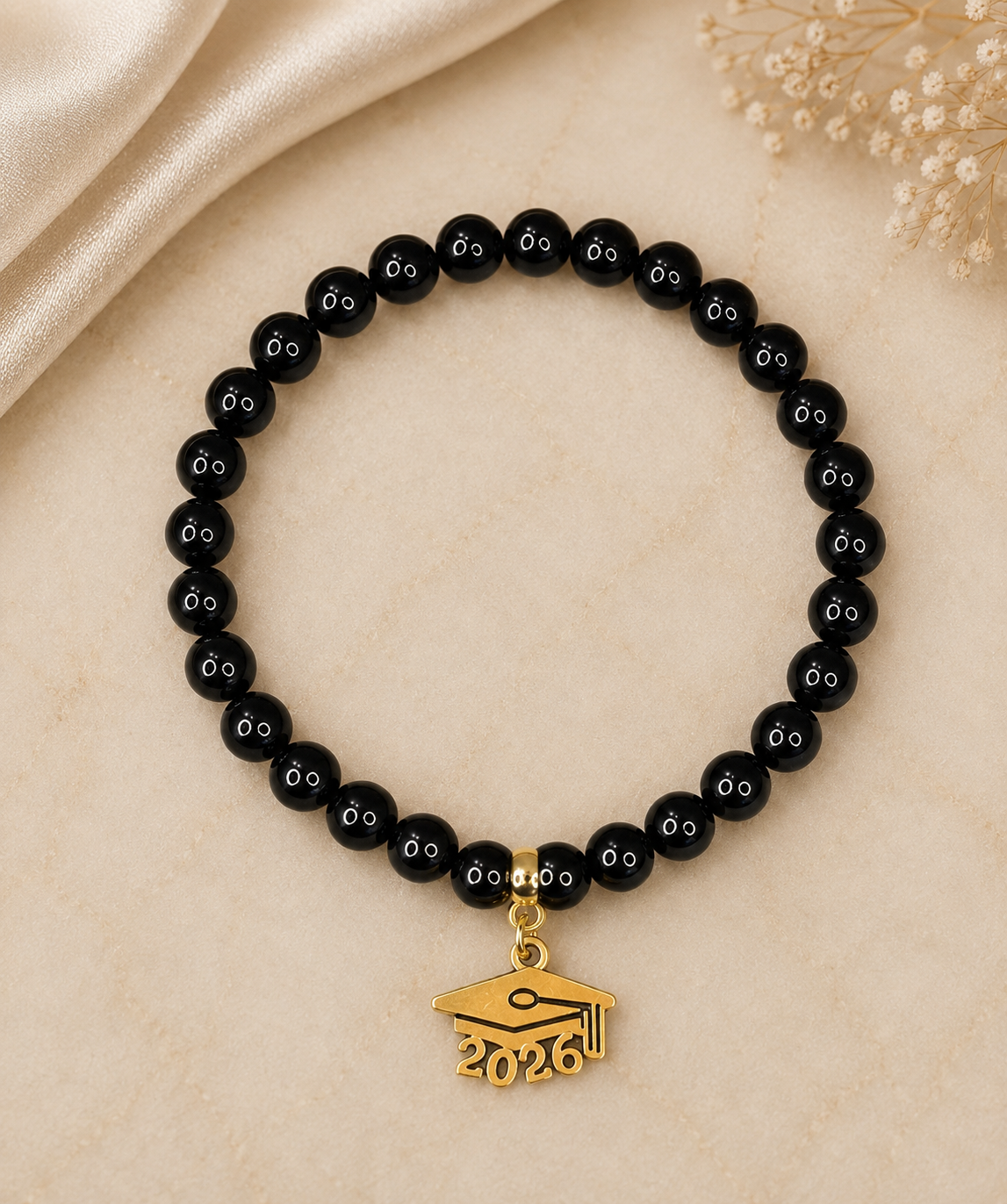 A beaded bracelet with glossy black round beads, each engraved with a small white 'oo' motif, and a gold-tone graduation cap charm featuring the year '2026'. The bracelet is displayed on a light beige textured surface with a satin fabric and dried flowers in the background.