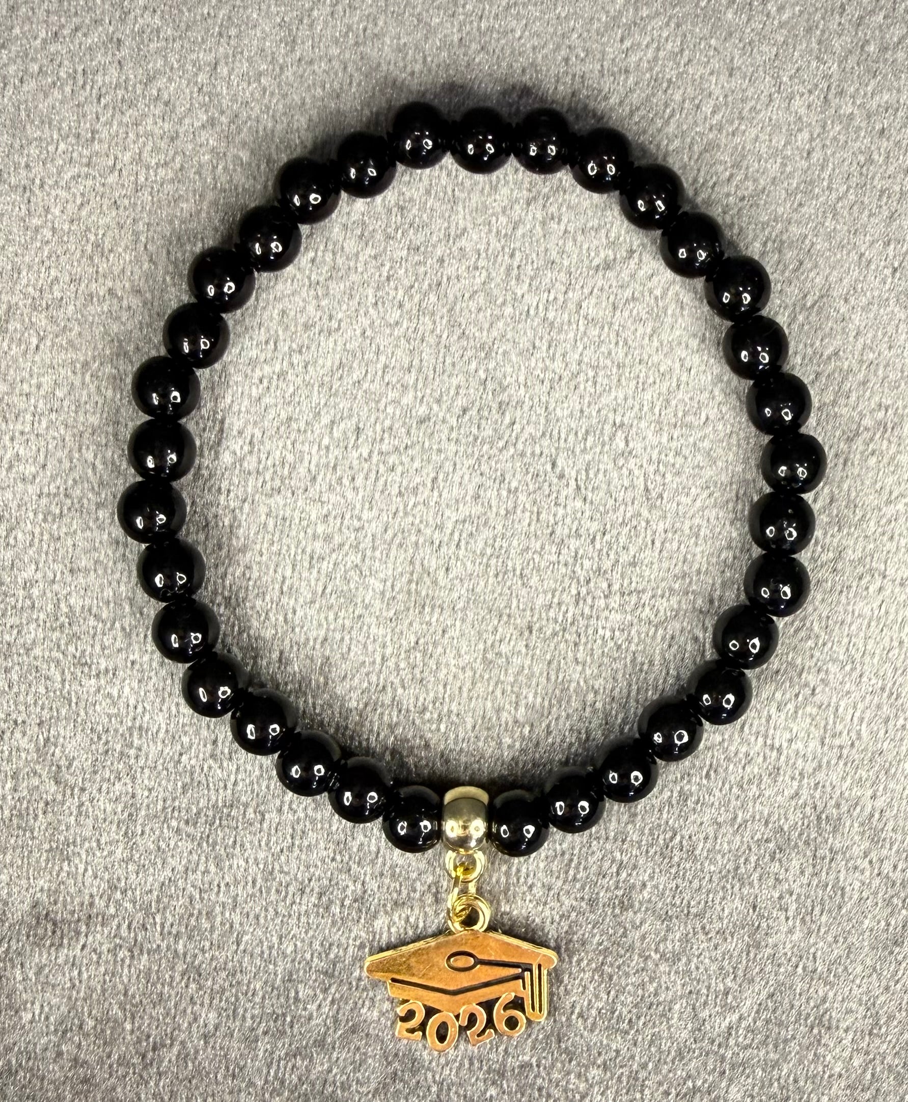A black beaded bracelet with a gold-colored graduation cap charm featuring the year '2026' engraved on it, displayed on a textured gray fabric background.