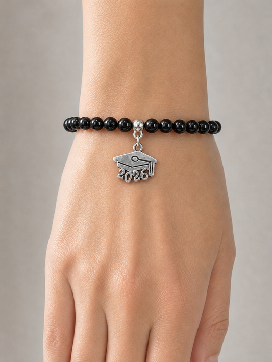 A black beaded bracelet worn on a person's wrist, featuring a silver charm shaped like a graduation cap with the year 2026 engraved on it, shown against a neutral gray background.