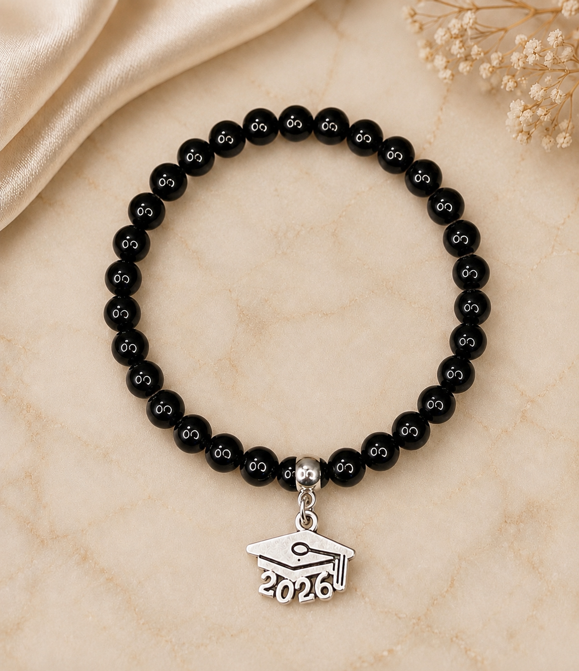 A beaded bracelet with glossy black round beads, each engraved with a small white semicolon, and a silver-toned graduation cap charm engraved with '2026', displayed on a beige marble surface with satin fabric and dried flowers.