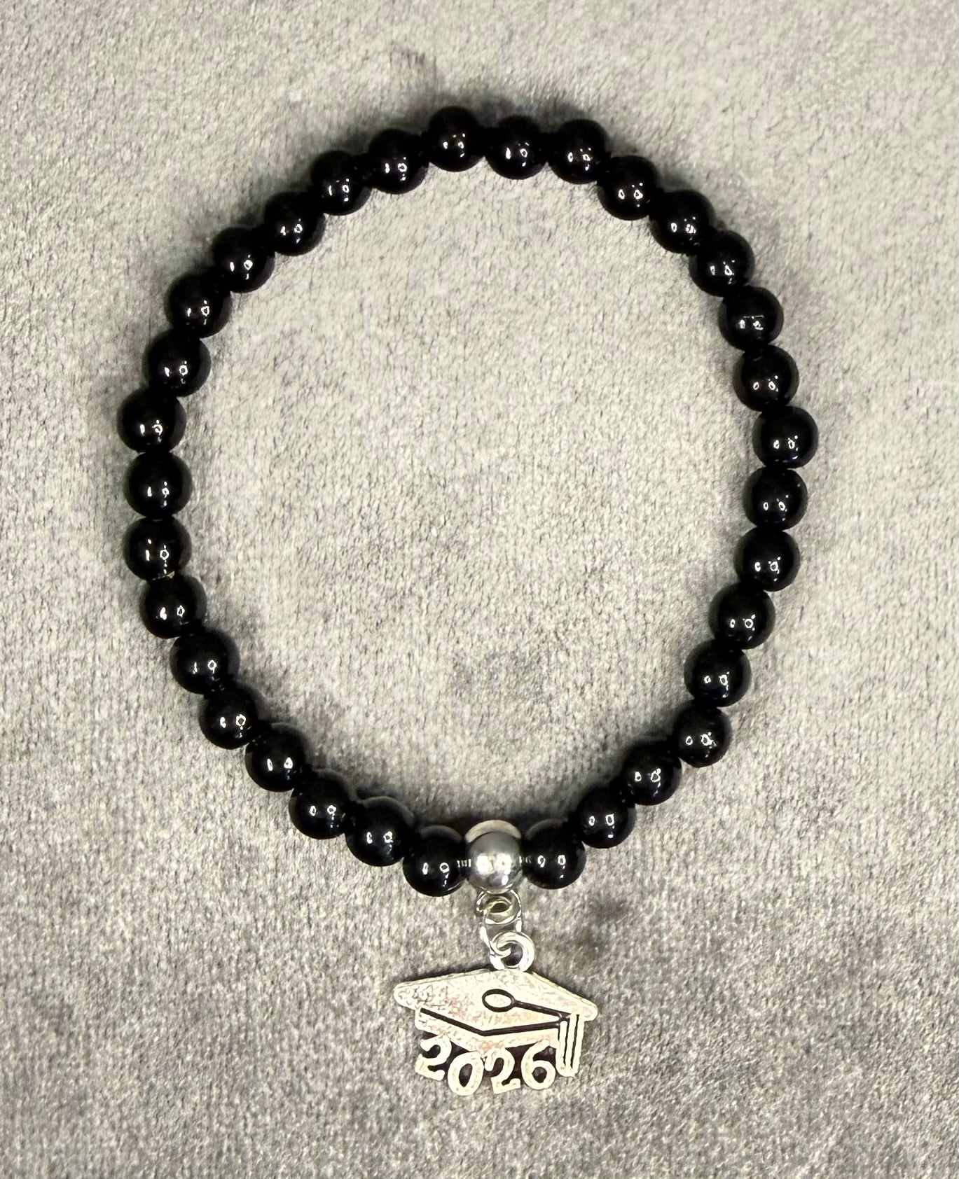 A beaded stretch bracelet made of glossy black round beads, featuring a silver-toned charm shaped like a graduation cap with the year 2026 engraved on it, displayed on a textured gray background.