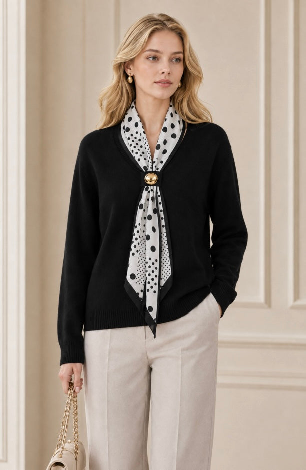 A woman models a black knit cardigan sweater with a black and white polka dot scarf tied around the neck, paired with beige trousers and holding a small beige handbag.