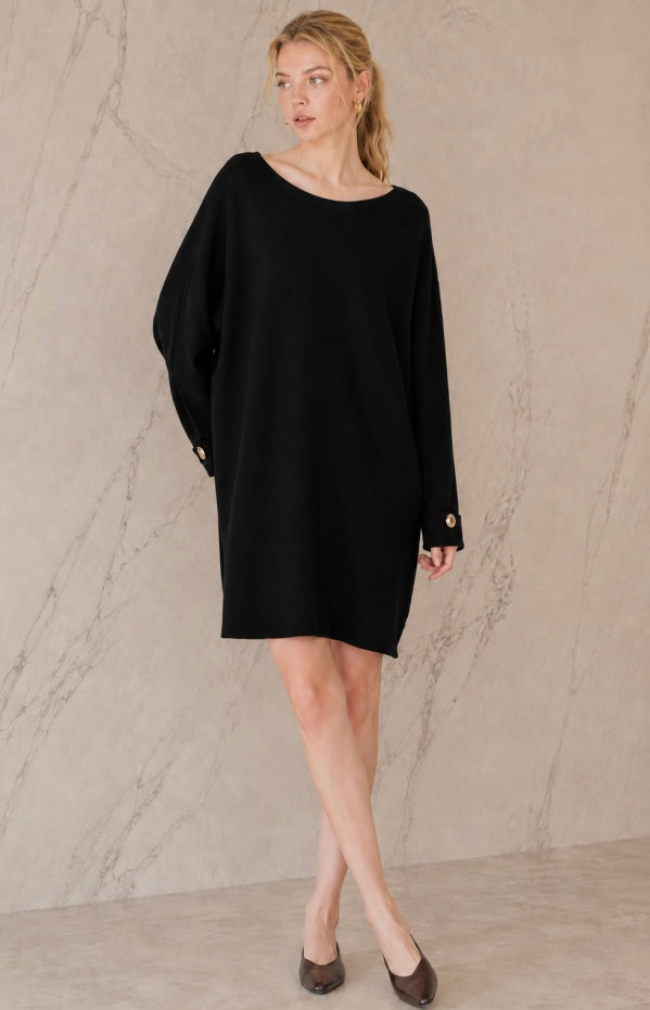A woman modeling a black oversized long-sleeve mini dress with a boat neckline and small gold button details on the cuffs, standing against a light beige marble-textured wall.