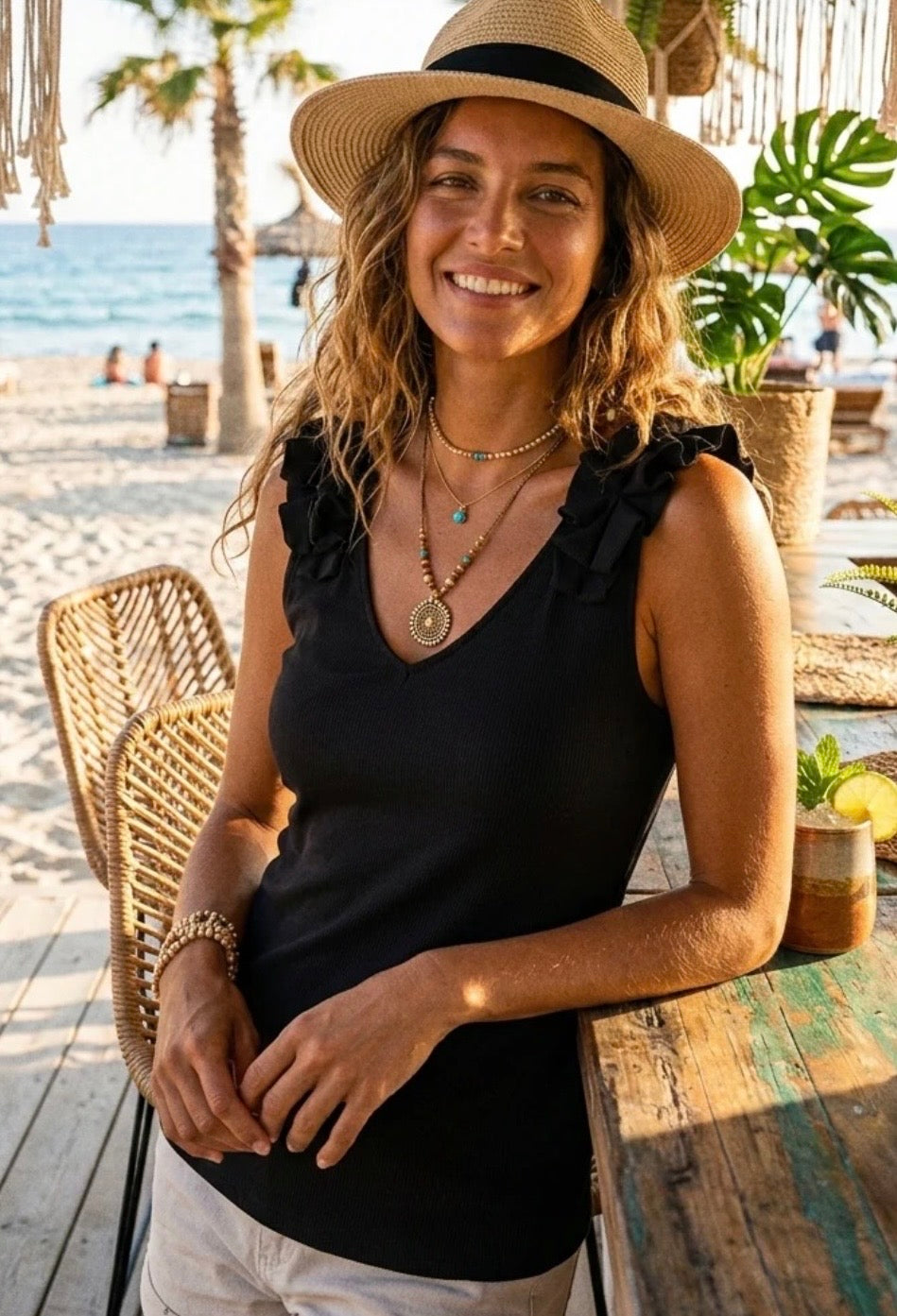 A woman wearing a black ruffle-sleeve V-neck top, a straw hat, and layered necklaces, leaning on a rustic table at a beachside cafe.