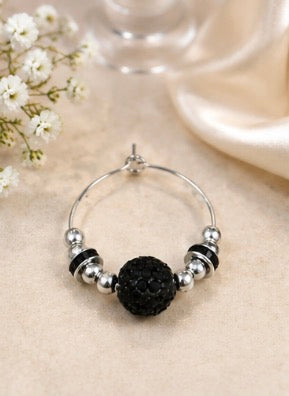 A silver wine glass charm with a central textured black bead and small metallic spacer beads, displayed on a beige fabric surface with small white flowers and a soft blurred background.