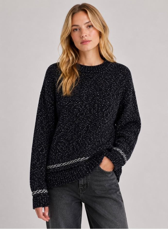 A woman wearing a black textured knit sweater with silver braided detailing on the cuffs and hem, paired with dark gray jeans, against a solid light gray background.