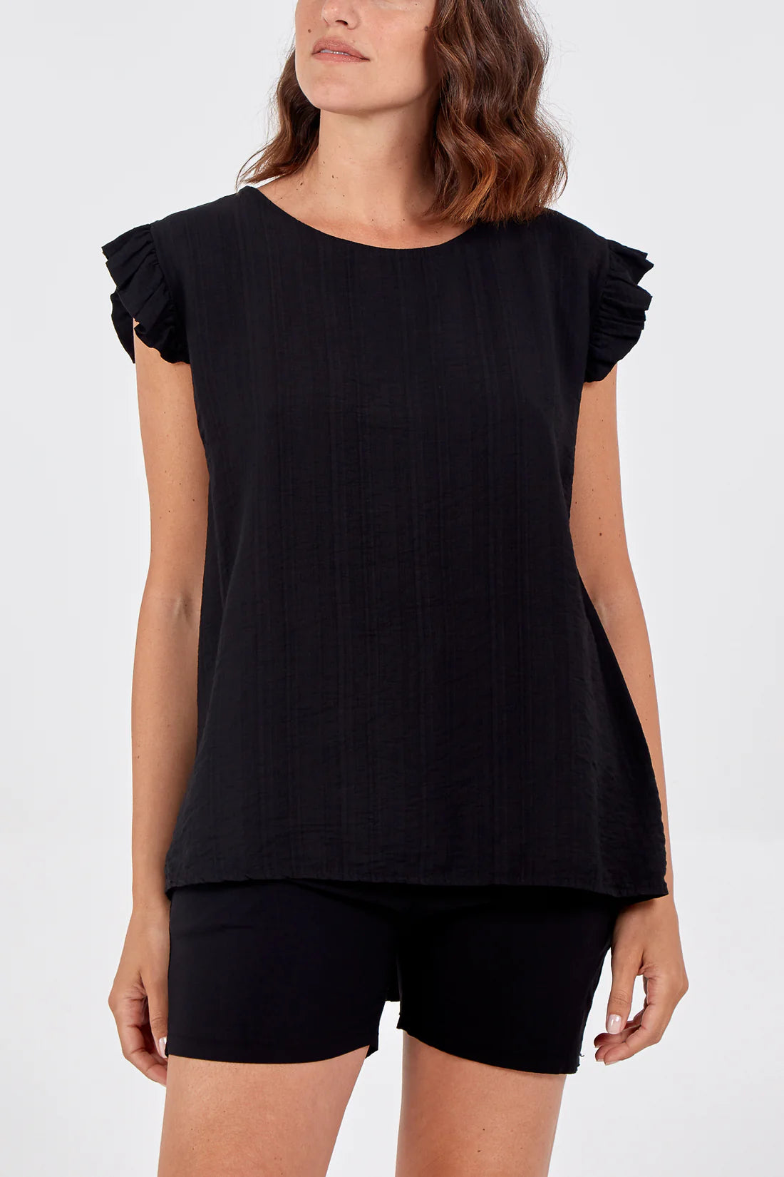 A woman wearing a black short-sleeve top with ruffled cap sleeves and black shorts, photographed against a plain light gray studio background.
