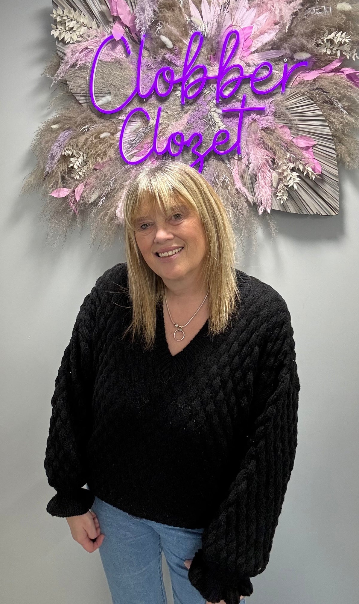 A woman with blonde hair wearing a chunky black cable-knit sweater and light blue jeans, standing in front of a gray wall with a decorative dried floral arrangement and purple neon sign that reads 'Clobber Closet'.