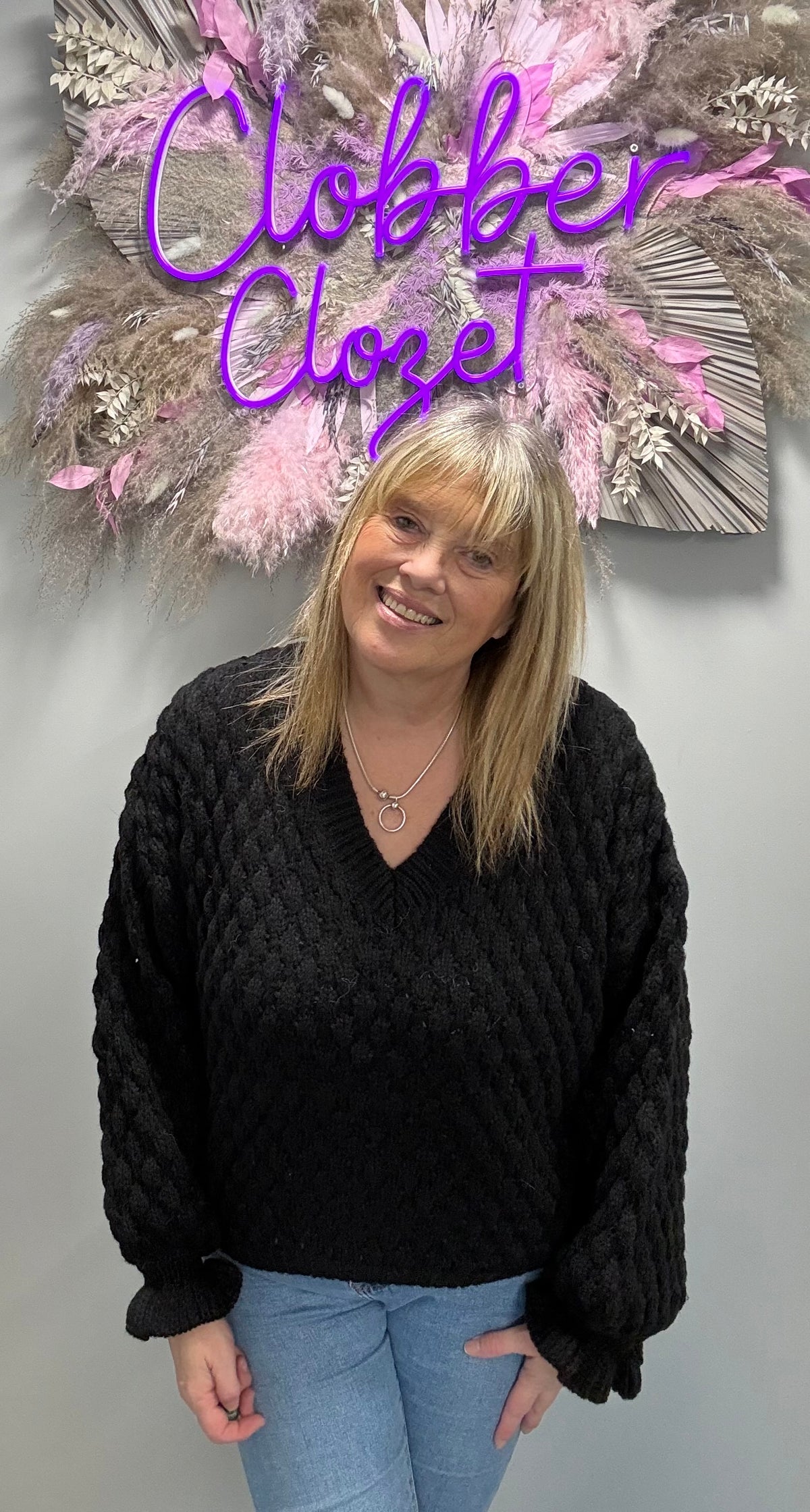 A woman wearing a black cable-knit sweater and light blue jeans stands in front of a gray wall with a decorative floral arrangement and a purple 'Clober Closet' sign behind her.