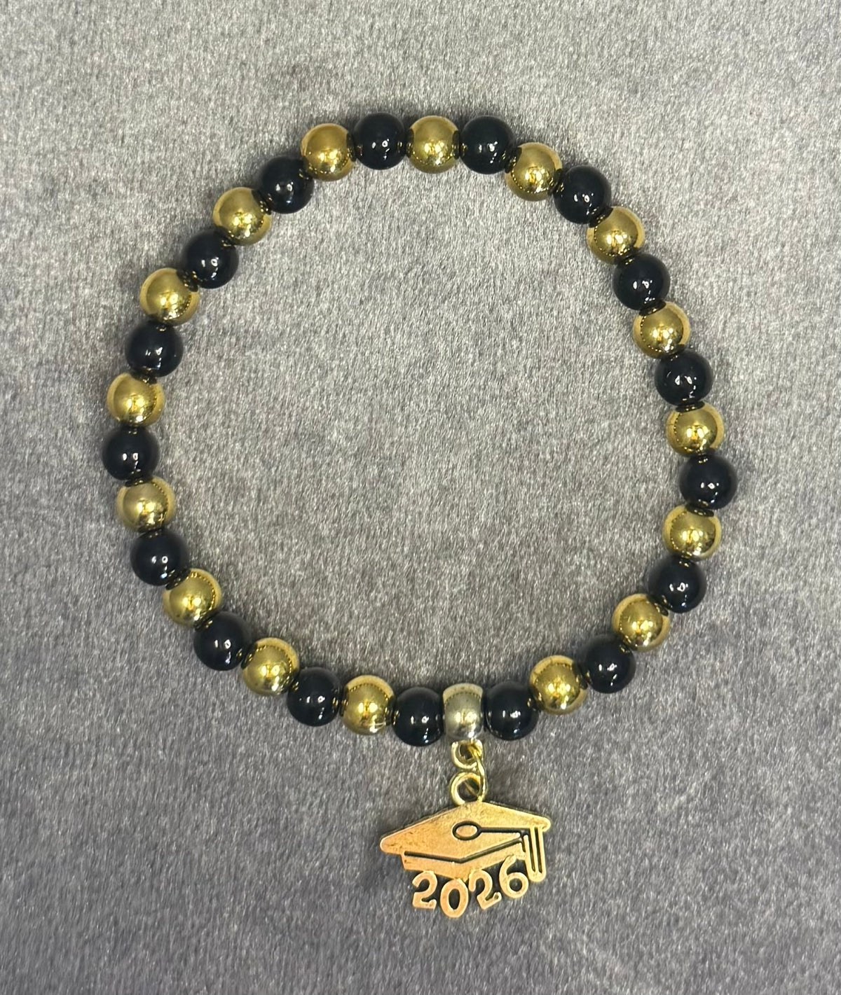 A beaded stretch bracelet featuring alternating glossy black and metallic gold beads, with a gold charm shaped like a graduation cap and the year '2026' engraved at the bottom, displayed on a textured gray background.