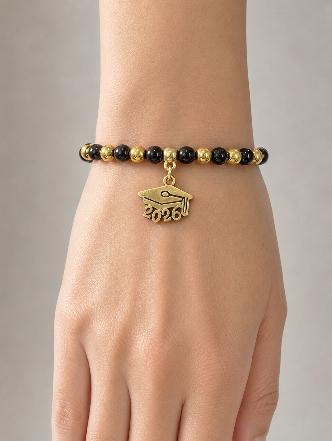 A gold and black beaded bracelet worn on a wrist, featuring a gold graduation cap charm with the year '2026' engraved on it, shown on a model's hand against a neutral gray background.