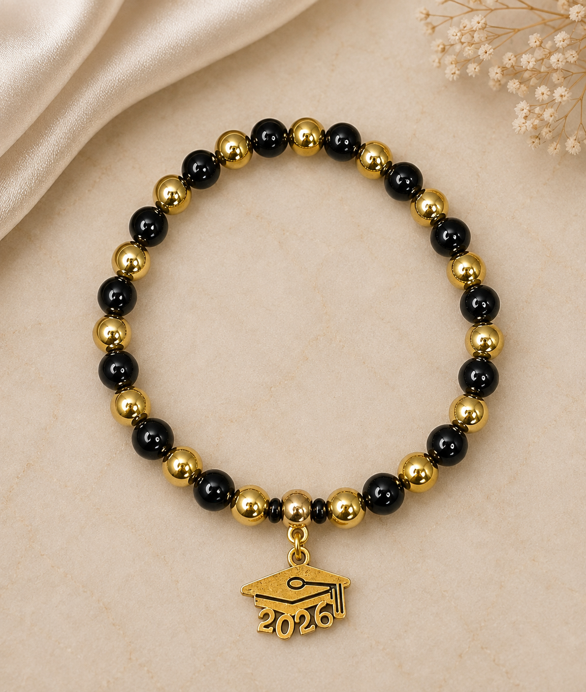 A beaded stretch bracelet featuring alternating glossy black and polished gold beads, with a small gold graduation cap charm engraved with '2026'. The bracelet is displayed on a light beige textured surface with a cream satin fabric and dried flowers in the background.