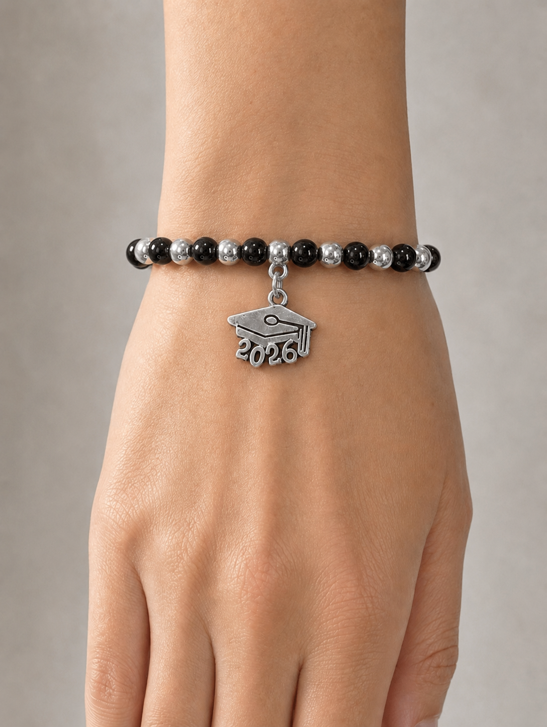 A close-up of a person's wrist wearing a beaded bracelet with black and silver beads and a silver graduation cap charm engraved with '2026'.