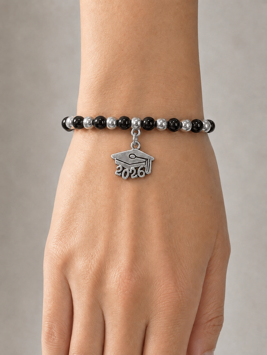 A close-up of a person's wrist wearing a beaded bracelet with black and silver beads and a silver graduation cap charm engraved with '2026'.